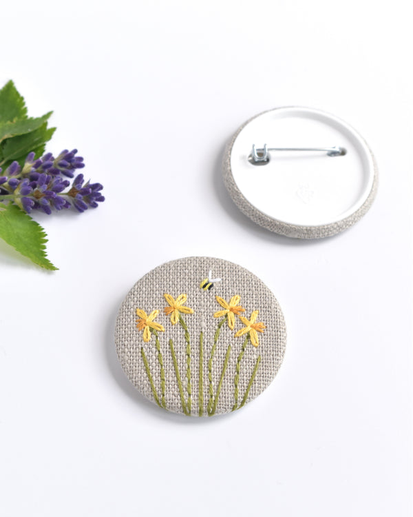 Embroidered daffodil pin badge. Four little yellow daffodil flowers and a tiny bumble bee are delicately hand sewn onto beige natural linen fabric, covering the front of the small round badge.
The round badge measures 45mm in diameter and comes attached to a branded white card.
Handmade by Stitch Galore
