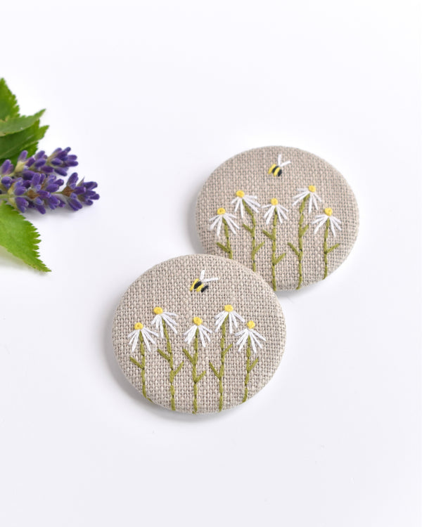 Embroidered daisy pin badge. Five little white daisy flowers and a tiny bumble bee are delicately hand sewn onto beige natural linen fabric, covering the front of the small round badge.
The round badge measures 45mm in diameter and comes attached to a branded white card.
Handmade by Stitch Galore
