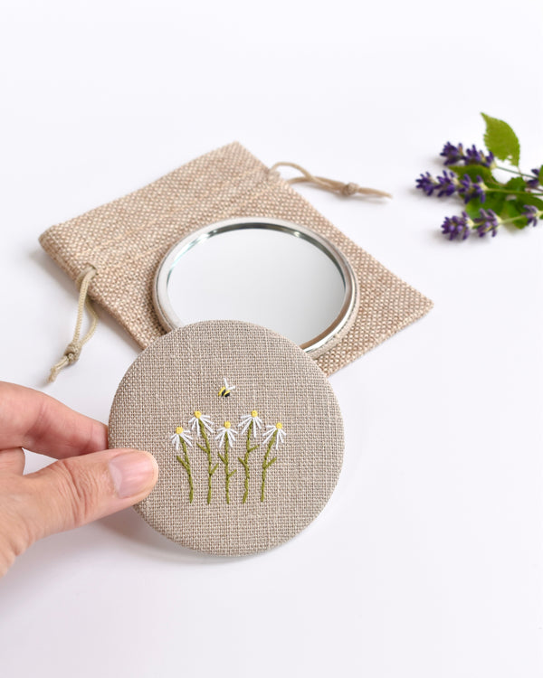 Embroidered daisy pocket mirror. Little white daisy flowers and a tiny bumble bee are delicately hand sewn onto natural linen fabric, covering the back of the small round hand mirror.
The round pocket mirror measures 77mm in diameter and comes with a linen drawstring pouch.
Image shows the pretty pocket mirror sitting on top of the linen pouch.
