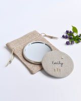 Embroidered personalised pocket mirror. An embroidered name, little flowers and a tiny bumble bee are delicately sewn onto natural linen fabric, covering the back of the small round hand mirror.
The round pocket mirror measures 77mm in diameter and comes with a linen drawstring pouch.
Image shows the pretty pocket mirror sitting on top of the linen pouch.
Handmade by Stitch Galore.
