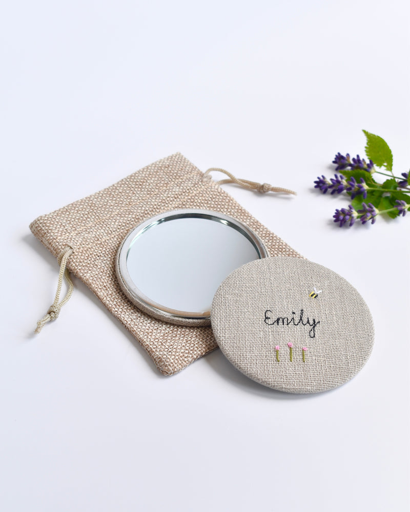 Embroidered personalised pocket mirror. An embroidered name, little flowers and a tiny bumble bee are delicately sewn onto natural linen fabric, covering the back of the small round hand mirror.
The round pocket mirror measures 77mm in diameter and comes with a linen drawstring pouch.
Image shows the pretty pocket mirror sitting on top of the linen pouch.
Handmade by Stitch Galore.
