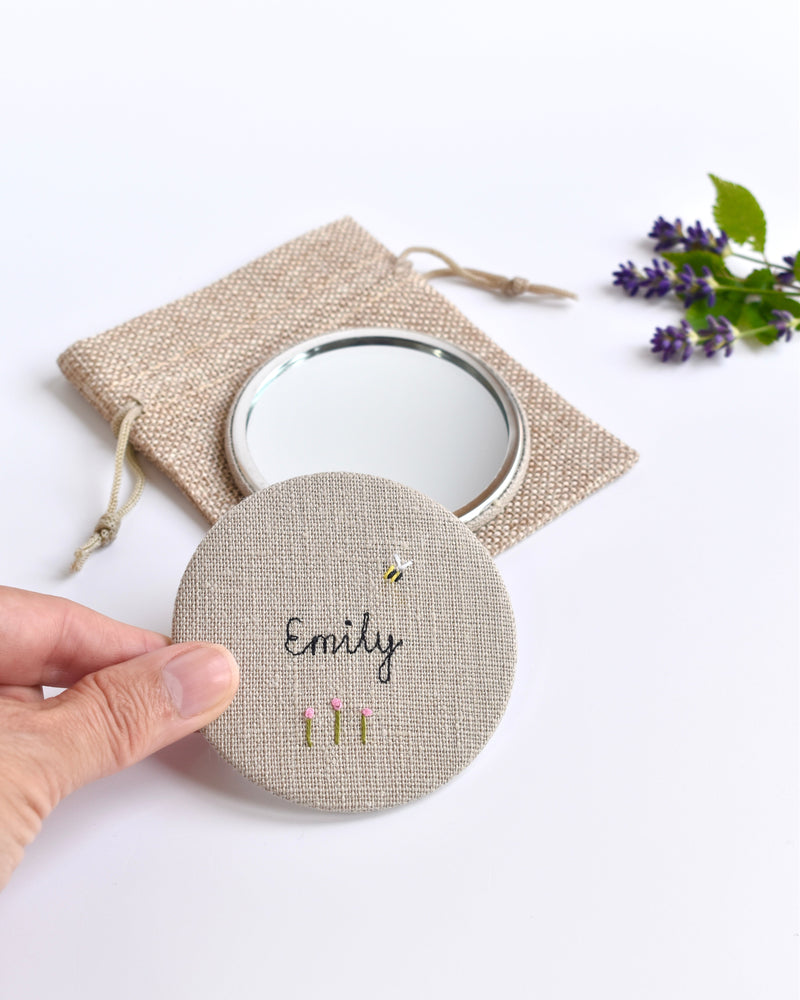 Embroidered personalised pocket mirror. An embroidered name, little flowers and a tiny bumble bee are delicately sewn onto natural linen fabric, covering the back of the small round hand mirror.
The round pocket mirror measures 77mm in diameter and comes with a linen drawstring pouch.
Image shows the pretty pocket mirror sitting on top of the linen pouch.
Handmade by Stitch Galore.
