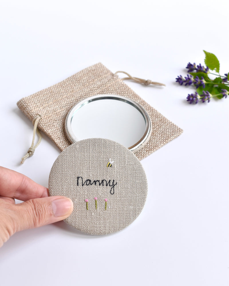 Embroidered personalised pocket mirror. An embroidered name, little flowers and a tiny bumble bee are delicately sewn onto natural linen fabric, covering the back of the small round hand mirror.
The round pocket mirror measures 77mm in diameter and comes with a linen drawstring pouch.
Image shows the pretty pocket mirror sitting on top of the linen pouch.
Handmade by Stitch Galore.
