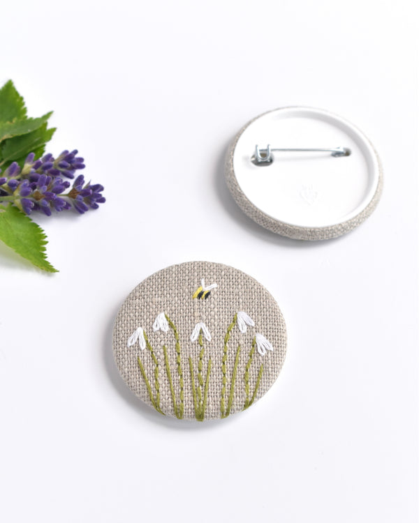 Embroidered snowdrop pin badge. Five little white snowdrop flowers and a tiny bumble bee are delicately hand sewn onto beige natural linen fabric, covering the front of the small round badge.
The round badge measures 45mm in diameter and comes attached to a branded white card.
Handmade by Stitch Galore
