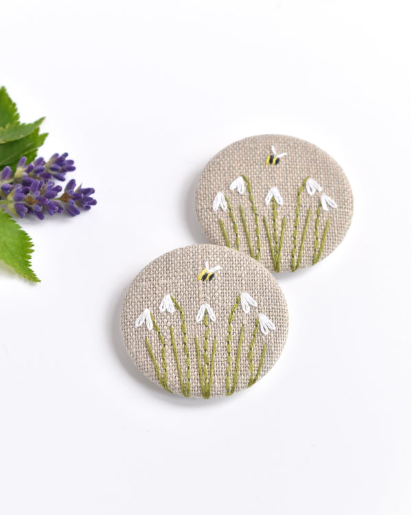 Embroidered snowdrop pin badge. Five little white snowdrop flowers and a tiny bumble bee are delicately hand sewn onto beige natural linen fabric, covering the front of the small round badge.
The round badge measures 45mm in diameter and comes attached to a branded white card.
Handmade by Stitch Galore
